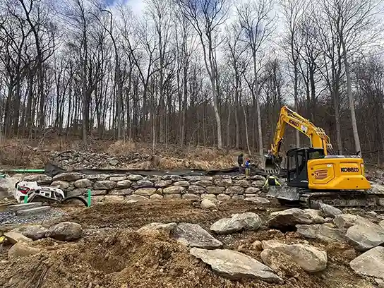 A yellow excavator is actively excavating a rock wall at a construction site.