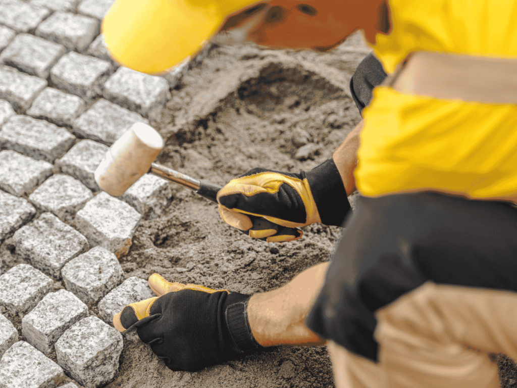 A man wearing a yellow shirt and hat is working on pavers installation on a sidewalk.