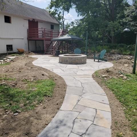 A stone pathway of pavers leads to a cozy fire pit, surrounded by greenery and inviting seating areas.